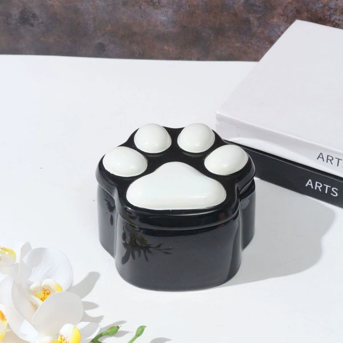 This pink and black paw ceramic pet urn features a stylish paw print lid, available in soft pink or sleek black, measuring approximately 12cm tall and 10cm wide. Crafted from durable ceramic, it offers secure storage for ashes of small to medium pets like cats or dogs, making it a heartfelt memorial display for North American pet owners on shelves or tables.