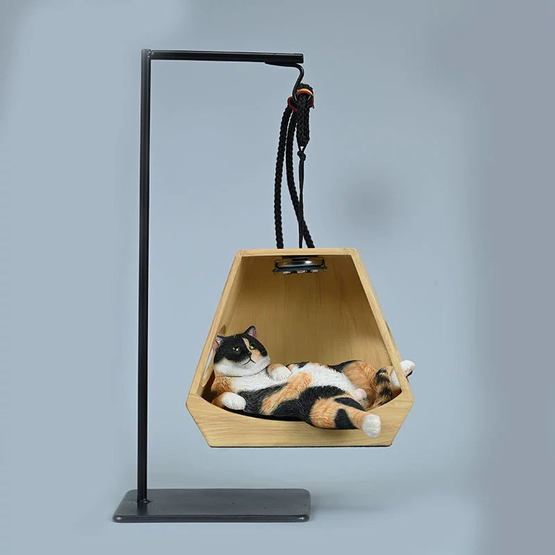 A whimsical decorative piece featuring a cat model lounging comfortably in a hanging basket shaped like a wooden hexagon. The basket is suspended from a metal frame with ropes, illuminated by a small overhead light, enhancing the cozy and serene appearance of the cat. This charming figurine adds a playful touch to any space.