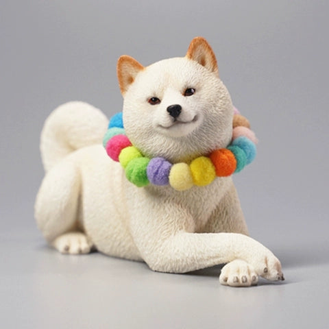 A collection of four Shiba Inu dog figurines in a prone position, each wearing a colorful pom-pom collar. The dogs, with diverse coat colors—white, tan, red, and black—are depicted lying down in a relaxed manner. The figurines are highly detailed, showcasing the breed's distinct features such as curled tails and alert expressions.