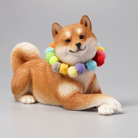 A collection of four Shiba Inu dog figurines in a prone position, each wearing a colorful pom-pom collar. The dogs, with diverse coat colors—white, tan, red, and black—are depicted lying down in a relaxed manner. The figurines are highly detailed, showcasing the breed's distinct features such as curled tails and alert expressions.