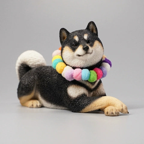 A collection of four Shiba Inu dog figurines in a prone position, each wearing a colorful pom-pom collar. The dogs, with diverse coat colors—white, tan, red, and black—are depicted lying down in a relaxed manner. The figurines are highly detailed, showcasing the breed's distinct features such as curled tails and alert expressions.