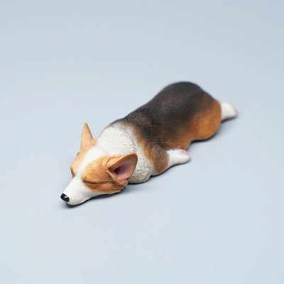 Two realistic 1/6 scale models of sleeping Corgi dogs. One model features a darker coat, while the other has a lighter coat, both capturing the adorable and relaxed pose of a sleeping Corgi.