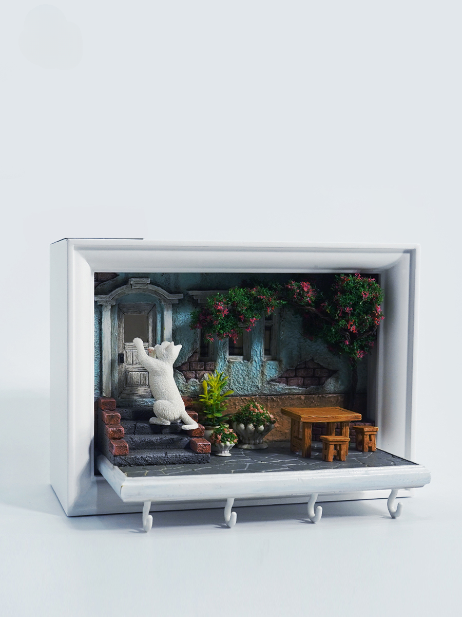 A detailed and charming art relief picture frame model featuring a white cat reaching up towards a window, surrounded by a beautifully crafted miniature garden with flowers, plants, and a rustic table with chairs. Perfect for adding a touch of whimsy and artistic flair to any room.