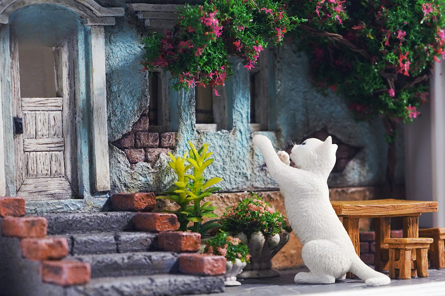 A detailed and charming art relief picture frame model featuring a white cat reaching up towards a window, surrounded by a beautifully crafted miniature garden with flowers, plants, and a rustic table with chairs. Perfect for adding a touch of whimsy and artistic flair to any room.