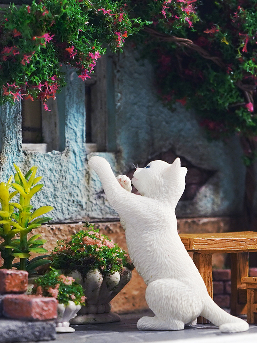 A detailed and charming art relief picture frame model featuring a white cat reaching up towards a window, surrounded by a beautifully crafted miniature garden with flowers, plants, and a rustic table with chairs. Perfect for adding a touch of whimsy and artistic flair to any room.