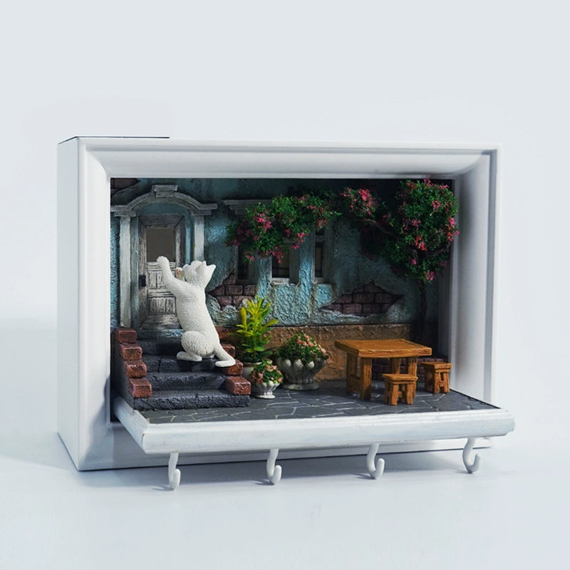 A detailed and charming art relief picture frame model featuring a white cat reaching up towards a window, surrounded by a beautifully crafted miniature garden with flowers, plants, and a rustic table with chairs. Perfect for adding a touch of whimsy and artistic flair to any room.