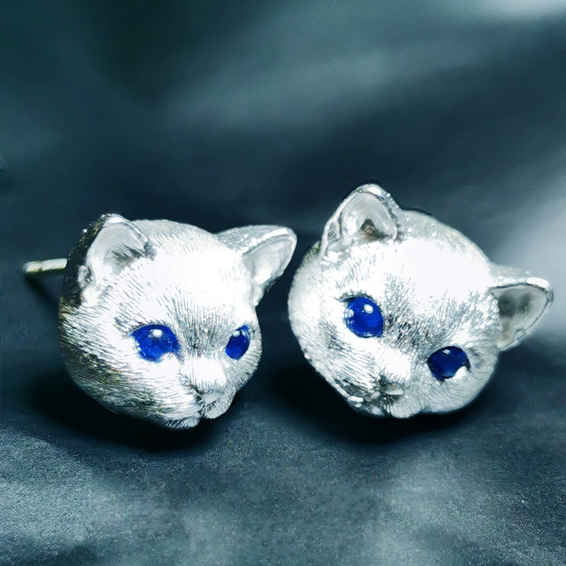 Close-up image of a handmade 925 sterling silver cat ring with blue gemstone eyes, highlighting the intricate details of the cat's face and the adorable tail wrapping around the finger. Matching earrings with similar detailing are also shown.