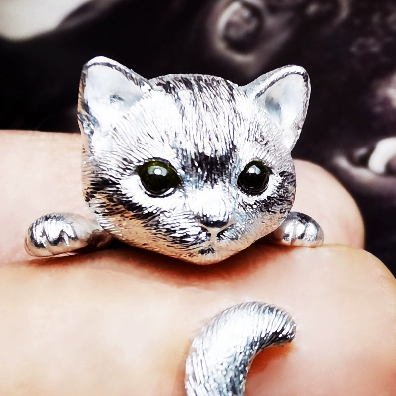 Close-up image of a handmade 925 sterling silver cat ring with blue gemstone eyes, highlighting the intricate details of the cat's face and the adorable tail wrapping around the finger. Matching earrings with similar detailing are also shown.