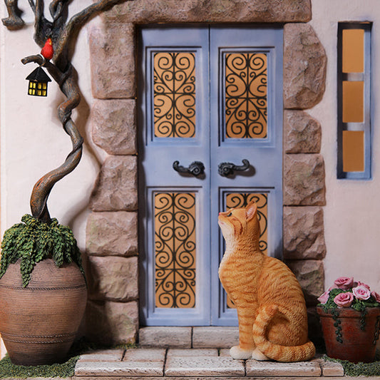 "A 1/6 scale model featuring an orange cat sitting in front of a detailed miniature blue door, adorned with intricate designs. The scene includes a potted plant and a small tree with a lantern hanging from it, capturing a whimsical and cozy setting."