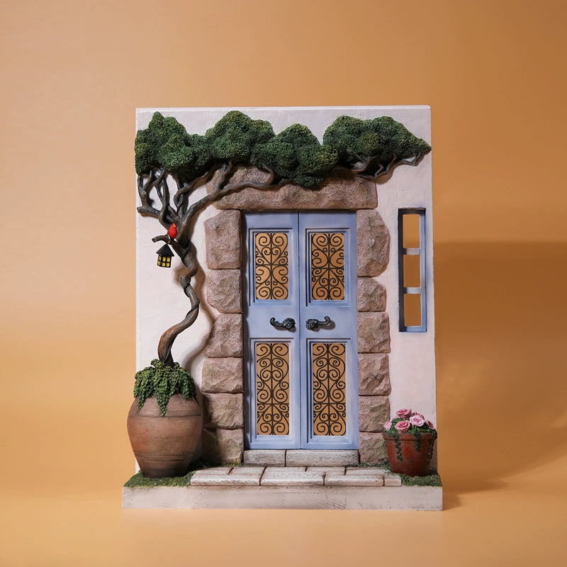 "A 1/6 scale model featuring an orange cat sitting in front of a detailed miniature blue door, adorned with intricate designs. The scene includes a potted plant and a small tree with a lantern hanging from it, capturing a whimsical and cozy setting."