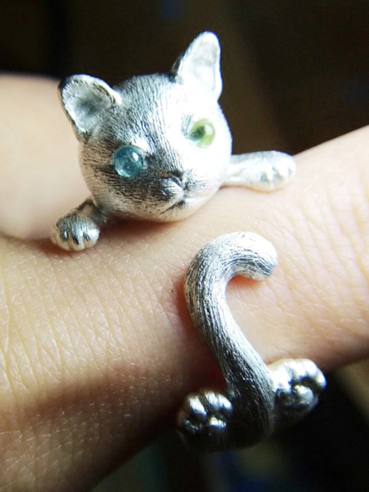 A handcrafted cat ring made of 925 sterling silver, featuring detailed fur textures and vibrant gemstone eyes, showcasing the playful pose of a kitten with its tail wrapped around the finger. Available in both silver and antique black finishes.