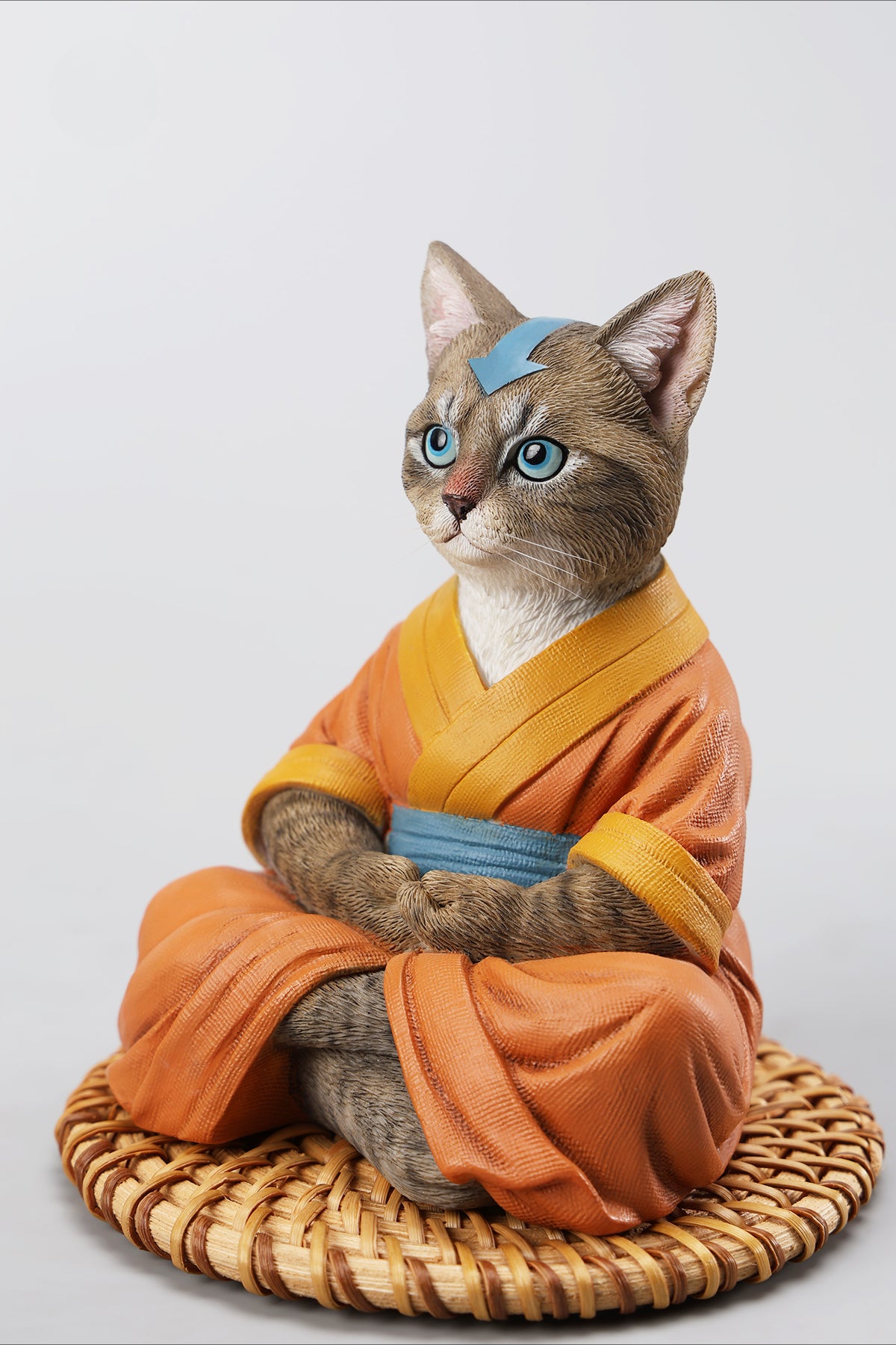 A handcrafted Airbender Cat Figurine in a meditative pose, dressed in an orange robe with a blue sash, sitting on a rattan-weaved cushion. This finely detailed resin sculpture captures the tranquility of mindfulness, perfect for cat lovers and meditation enthusiasts. An ideal collectible and decorative accent for any serene space.