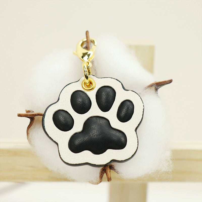 A collection of four handcrafted leather paw-shaped pendants in various colors, including black, green, tan, and white. Each pendant features a contrasting paw print, highlighting the high-quality craftsmanship and customizable options available. Ideal for pet owners who appreciate unique and personalized accessories.