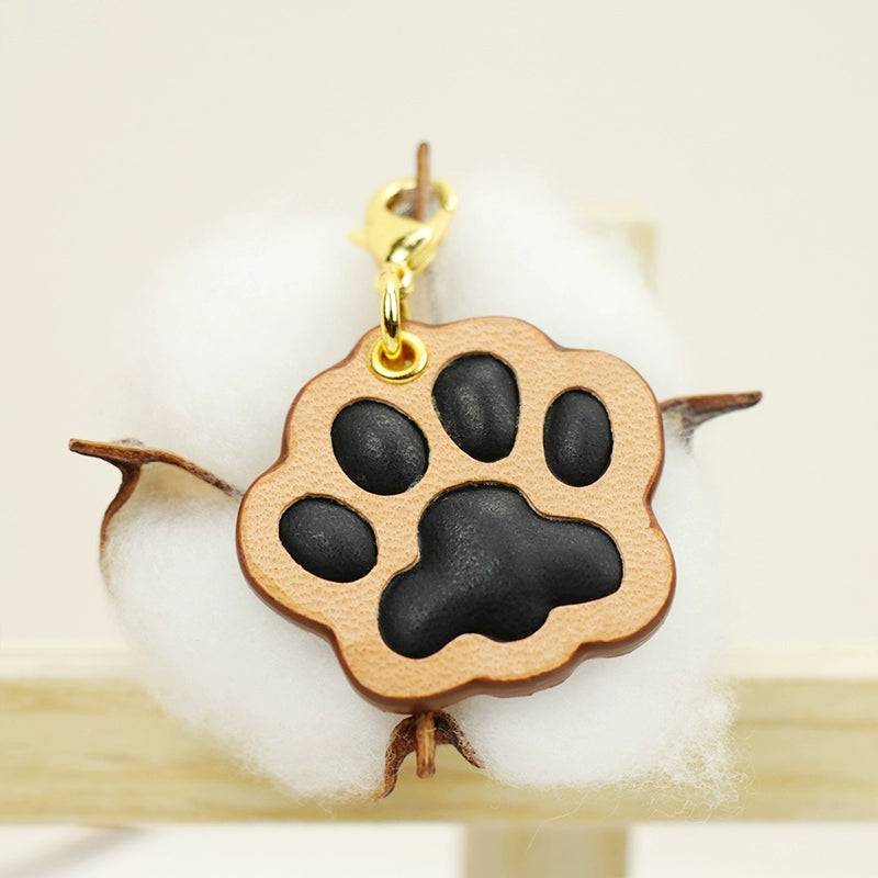 A collection of four handcrafted leather paw-shaped pendants in various colors, including black, green, tan, and white. Each pendant features a contrasting paw print, highlighting the high-quality craftsmanship and customizable options available. Ideal for pet owners who appreciate unique and personalized accessories.