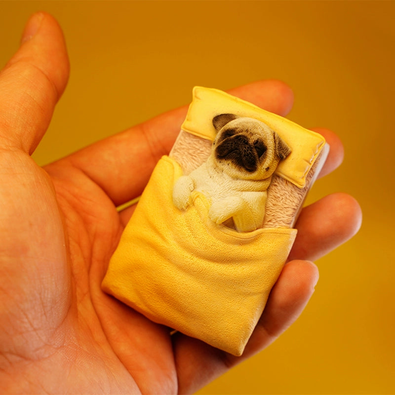 A cute pug figurine lies under a yellow quilt on a miniature bed, bathed in warm sunlight. The detailed craftsmanship showcases the pug's peaceful sleeping position with its paws tucked under the quilt, capturing a cozy and serene moment. Perfect for dog lovers and collectors.