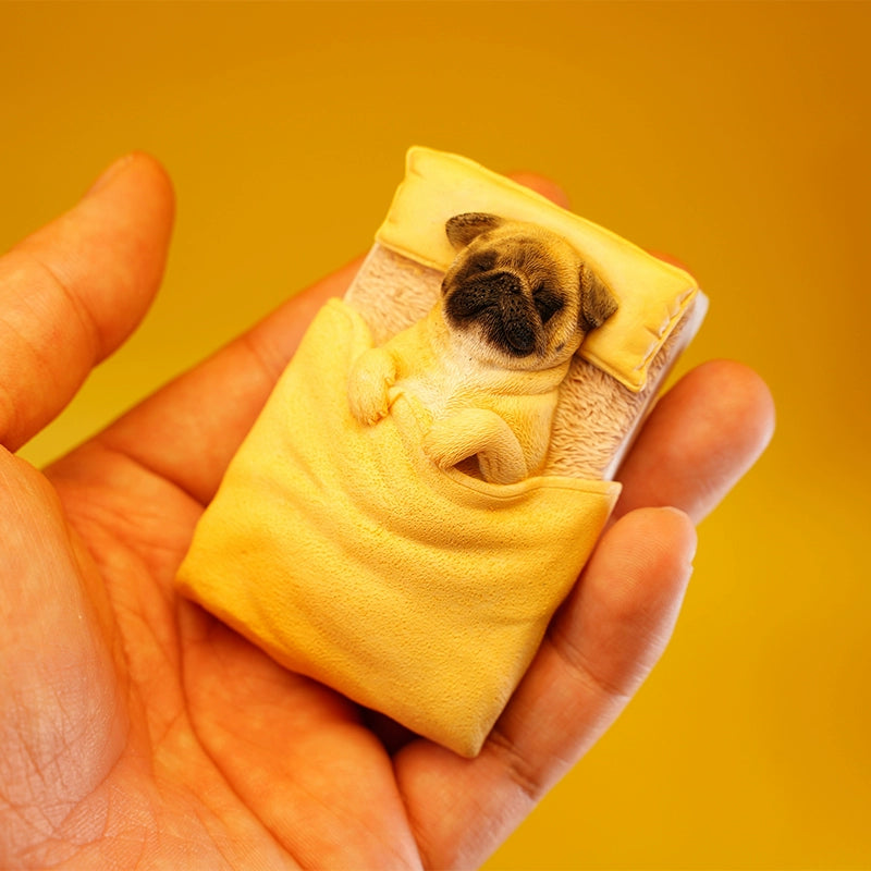 A cute pug figurine lies under a yellow quilt on a miniature bed, bathed in warm sunlight. The detailed craftsmanship showcases the pug's peaceful sleeping position with its paws tucked under the quilt, capturing a cozy and serene moment. Perfect for dog lovers and collectors.