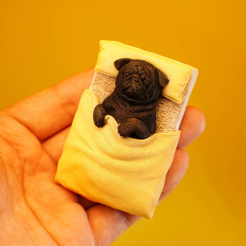 A cute pug figurine lies under a yellow quilt on a miniature bed, bathed in warm sunlight. The detailed craftsmanship showcases the pug's peaceful sleeping position with its paws tucked under the quilt, capturing a cozy and serene moment. Perfect for dog lovers and collectors.