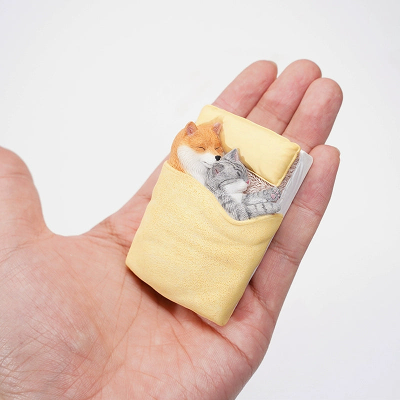 A charming figurine featuring a Shiba Inu dog snuggling with a cat under a yellow blanket. Both the dog and the cat are peacefully sleeping on a small bed, with detailed and realistic features that capture a heartwarming moment of companionship and comfort.