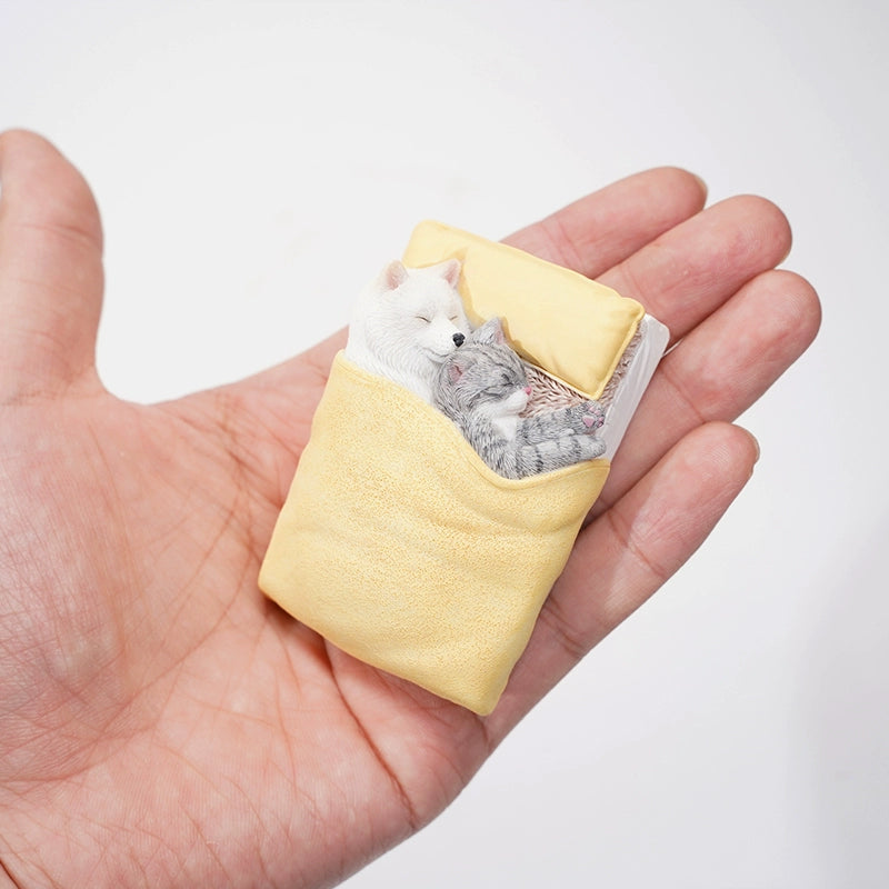 A charming figurine featuring a Shiba Inu dog snuggling with a cat under a yellow blanket. Both the dog and the cat are peacefully sleeping on a small bed, with detailed and realistic features that capture a heartwarming moment of companionship and comfort.