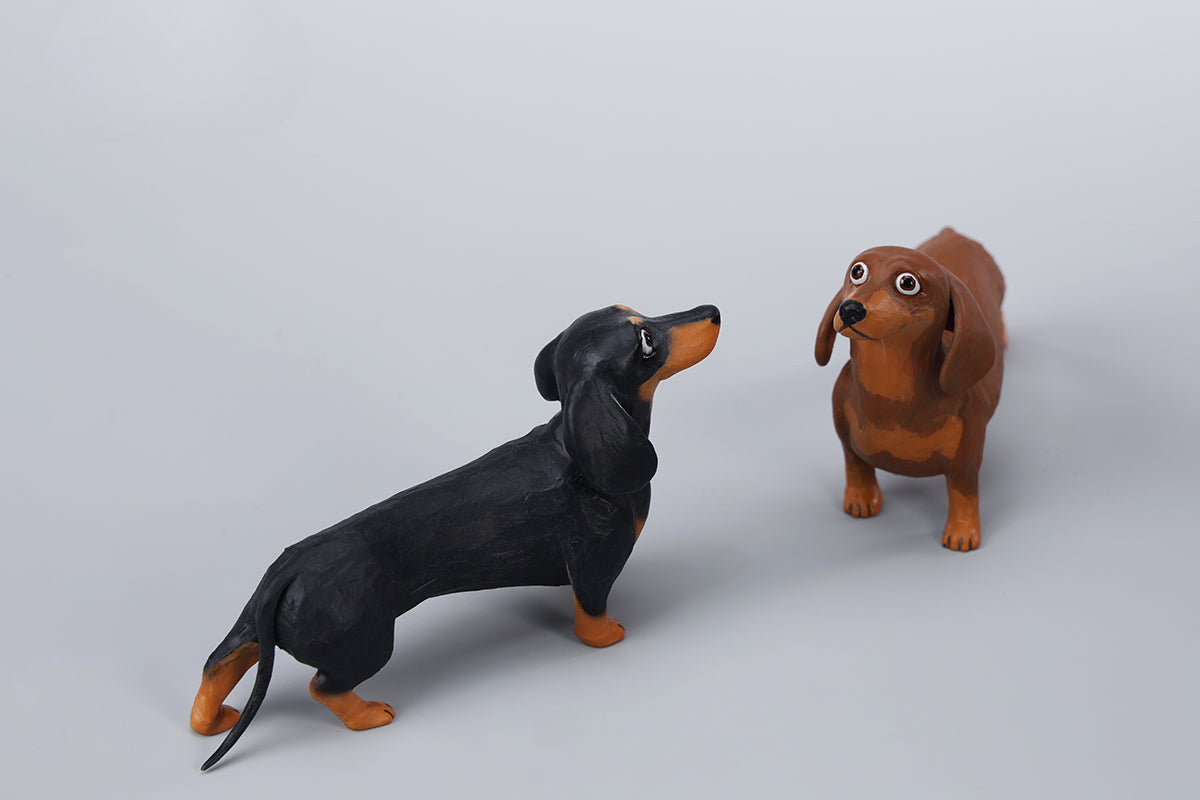 Two small clay Dachshund dog models, one black and one brown, captured in a realistic and lifelike manner. The models showcase detailed features, including expressive eyes and realistic fur textures. Perfect for collectors and dog enthusiasts.