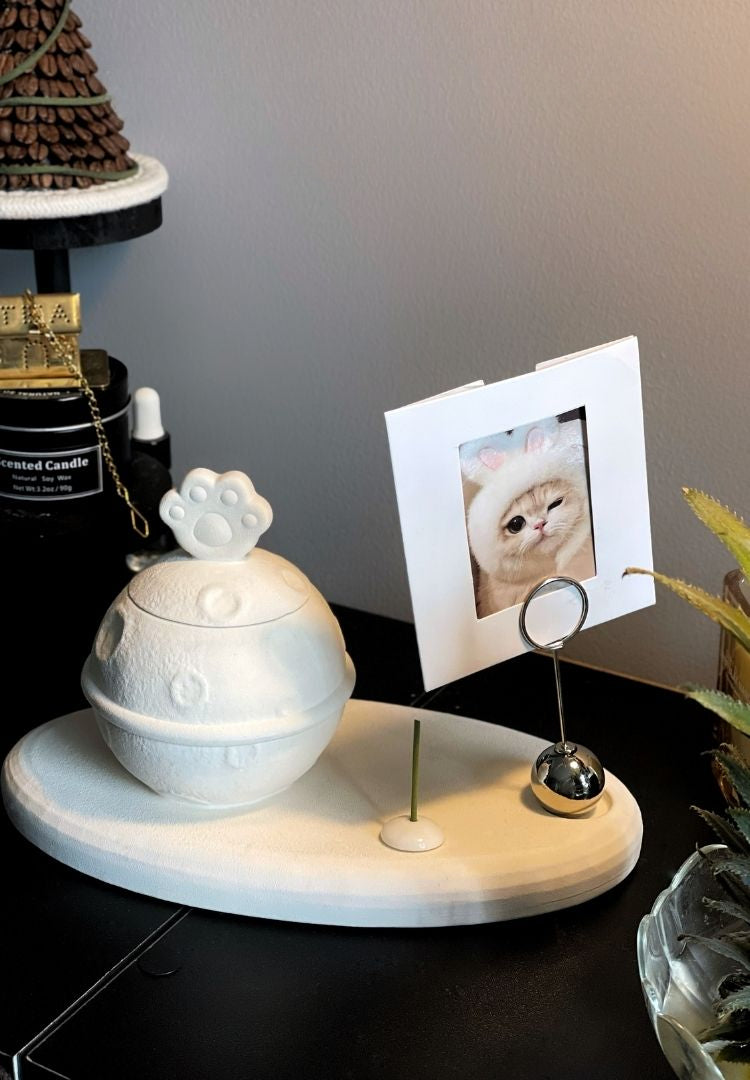 This white moon paw ceramic pet urn features a unique moon-shaped design with a paw print lid and detachable photo frame, measuring 22cm wide and 8cm tall with a 1-2cm base, holding 250ml of ashes. Perfect for North American pet owners honoring small pets like cats or dogs, it offers secure storage and a personalized memorial display for shelves or tables.