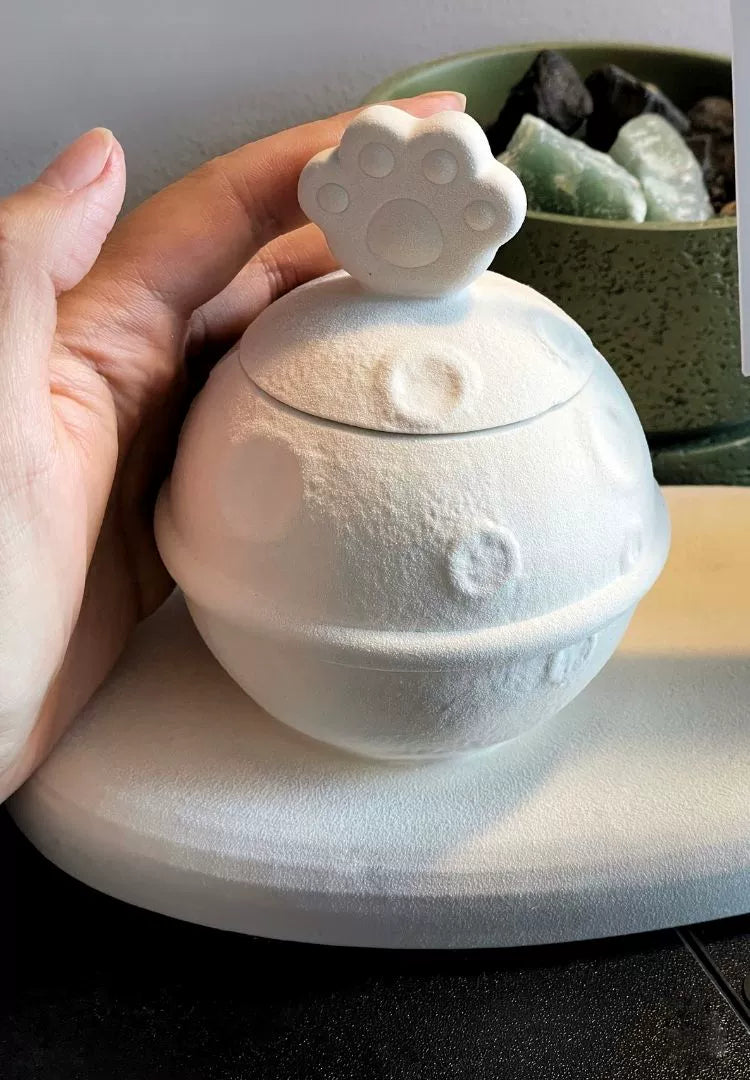 This white moon paw ceramic pet urn features a unique moon-shaped design with a paw print lid and detachable photo frame, measuring 22cm wide and 8cm tall with a 1-2cm base, holding 250ml of ashes. Perfect for North American pet owners honoring small pets like cats or dogs, it offers secure storage and a personalized memorial display for shelves or tables.