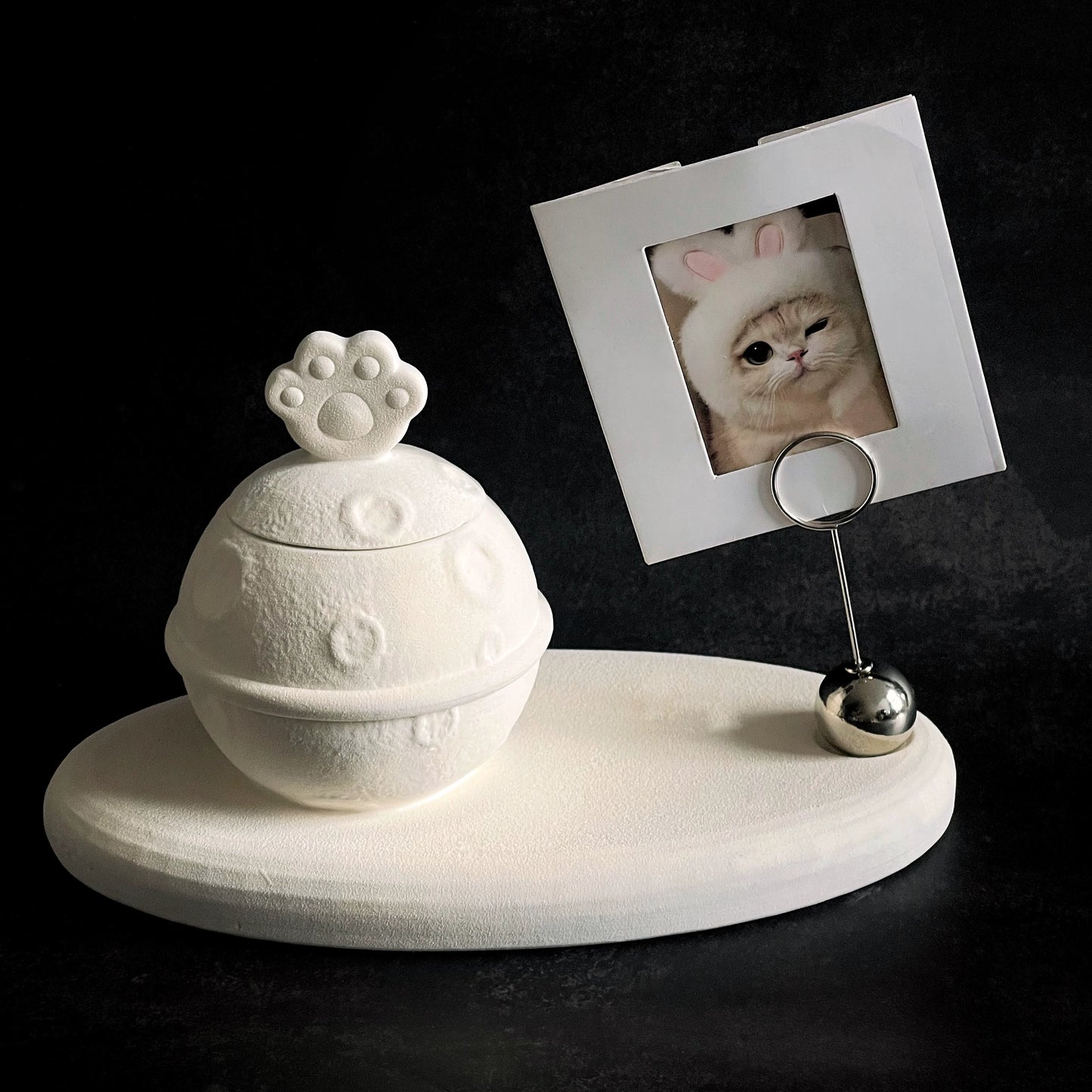 This white moon paw ceramic pet urn features a unique moon-shaped design with a paw print lid and detachable photo frame, measuring 22cm wide and 8cm tall with a 1-2cm base, holding 250ml of ashes. Perfect for North American pet owners honoring small pets like cats or dogs, it offers secure storage and a personalized memorial display for shelves or tables.