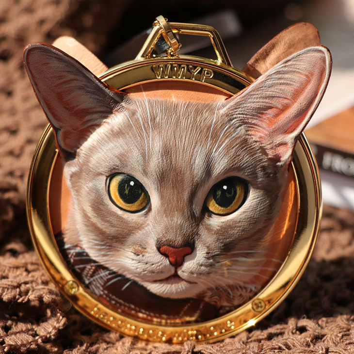 A striking leather carving of a curious-looking cat with large, expressive eyes, perfectly captured in a circular metal frame. The artist's attention to detail is evident in the cat's lifelike expression and the realistic texture of its fur. This handcrafted piece offers a unique and personal way to commemorate a beloved pet.